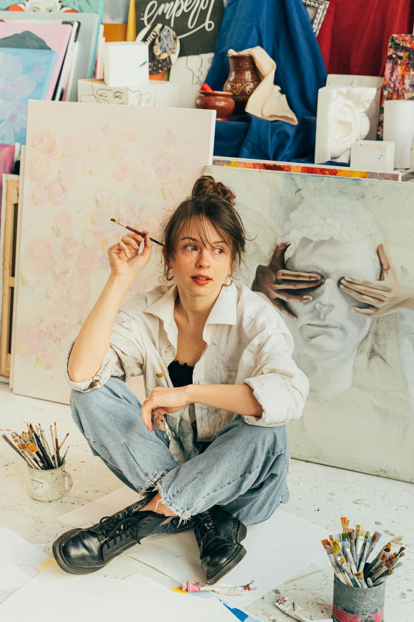 A young woman artist sits on the floor of a colorful studio surrounded by her artwork and supplies.