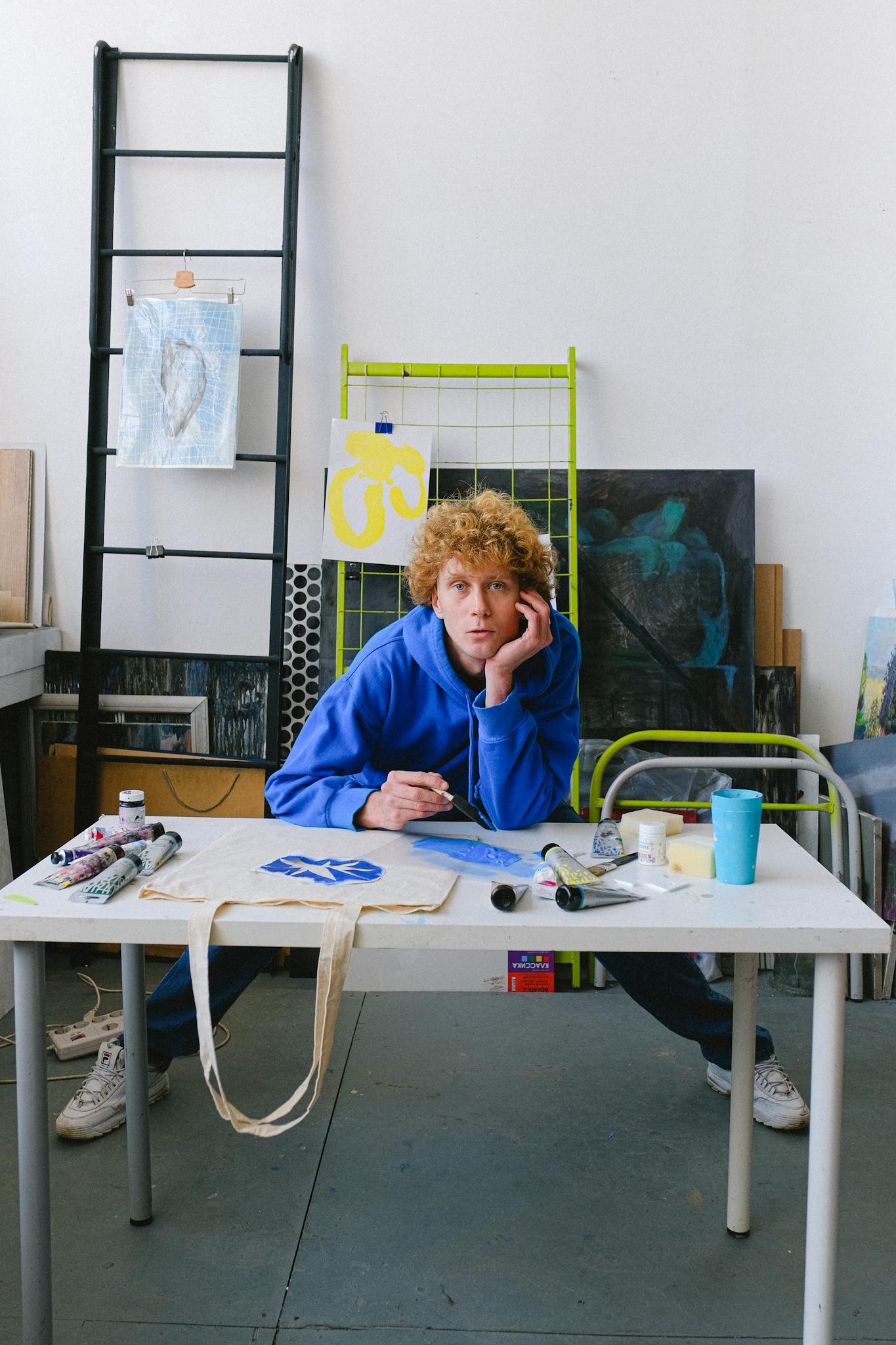 Full body of calm male designer looking at camera and painting while standing at table with spread legs in studio
