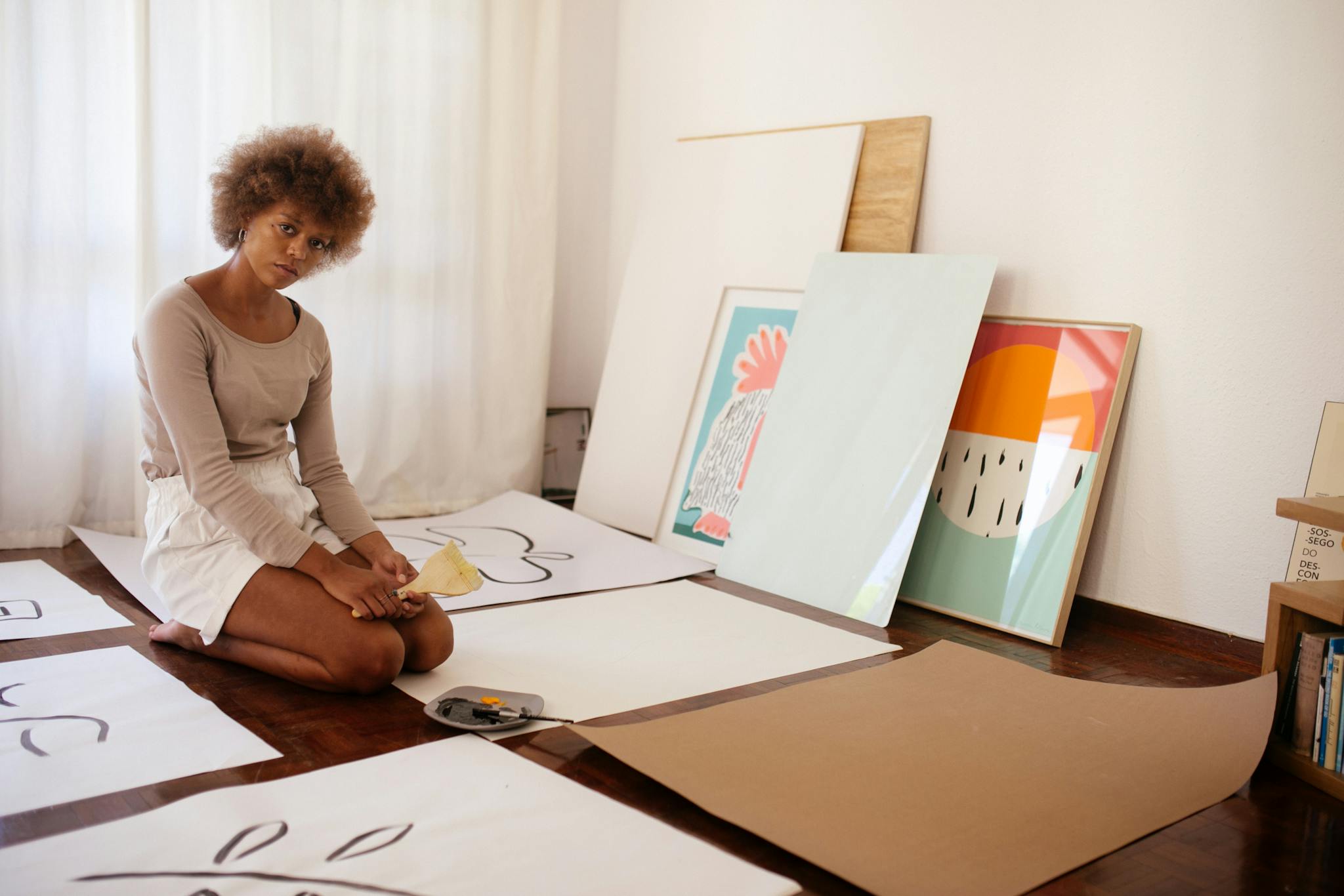Young woman artist sitting among canvases, thoughtfully creating art in her cozy home studio.